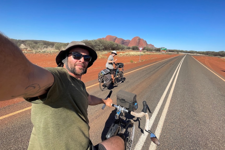 A man wearing sun glasses takes a selfie with his son, the road and Kata Tjuta in the background.