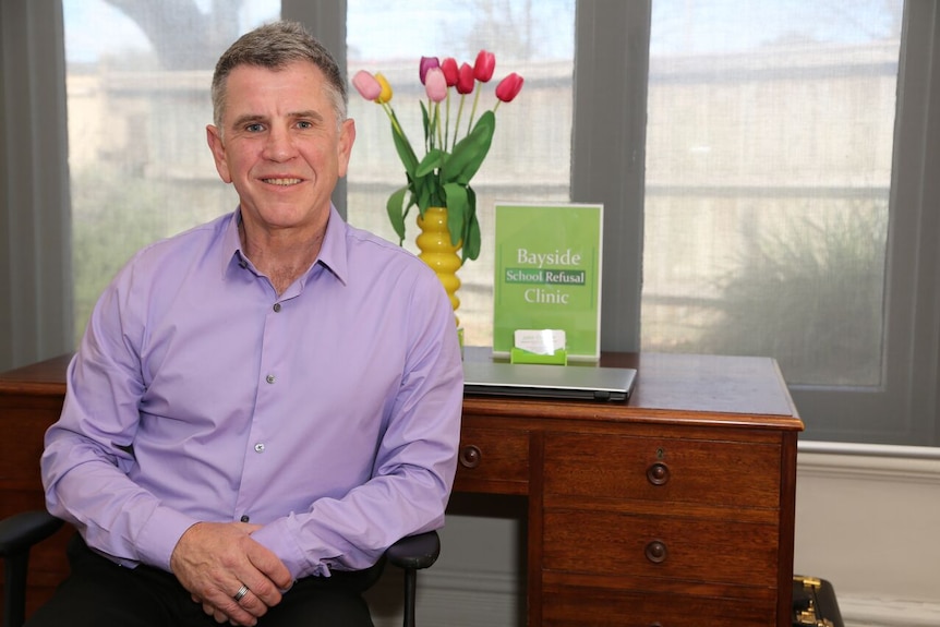 A middle aged man sits in front of a desk in a medical clinic.