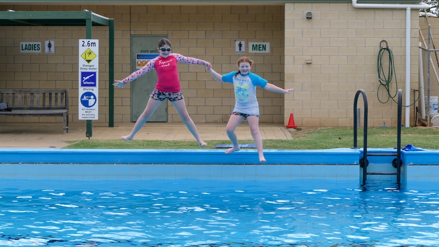 two primary school aged girls, sitting in bathers beside a pool with a sign which says 'Save Our Pool'