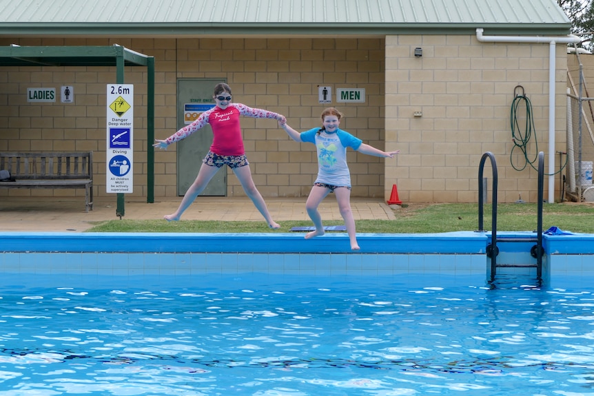 two primary school aged girls, sitting in bathers beside a pool with a sign which says 'Save Our Pool'