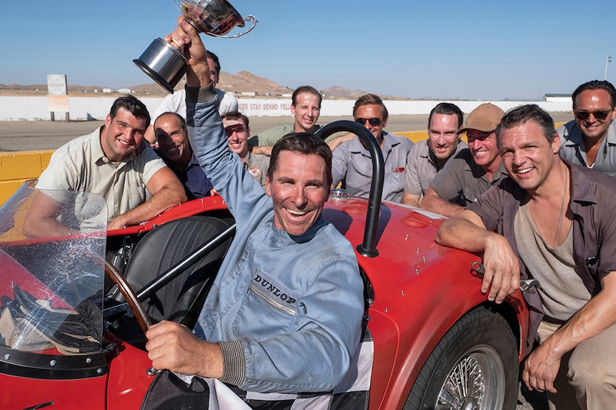 Christian Bale sits in race car and holds up trophy while surrounded by group of male mechanics on race track.