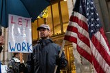 A man holds a sign which says "let us work" and holds an American flag.
