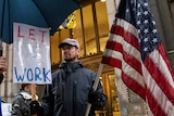 A man holds a sign which says "let us work" and holds an American flag.