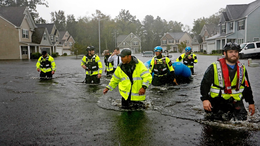 A group of men in high-vis work wear wade through a flooded street with one pulling along a blue inflatable raft