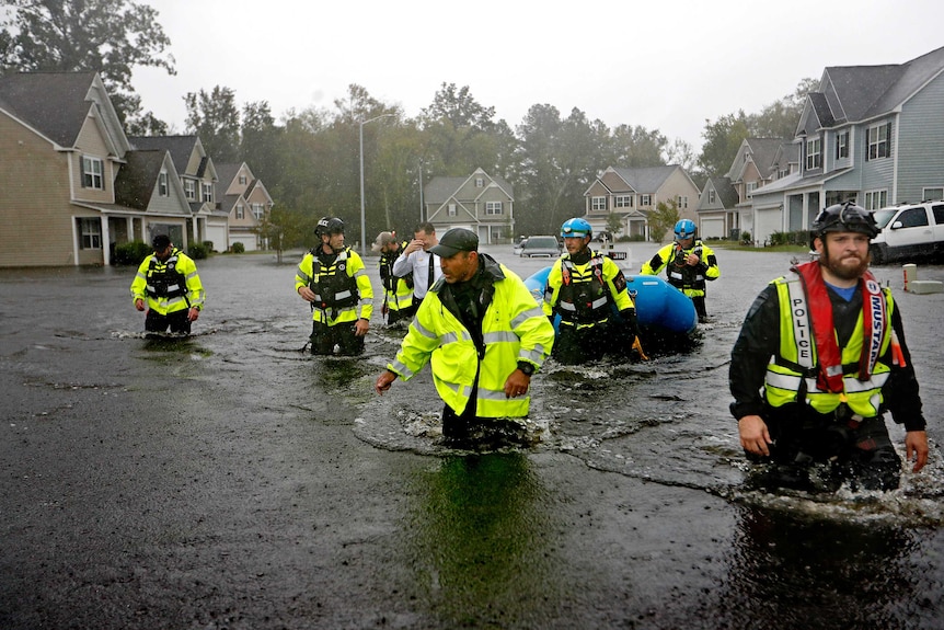 A group of men in high-vis work wear wade through a flooded street with one pulling along a blue inflatable raft