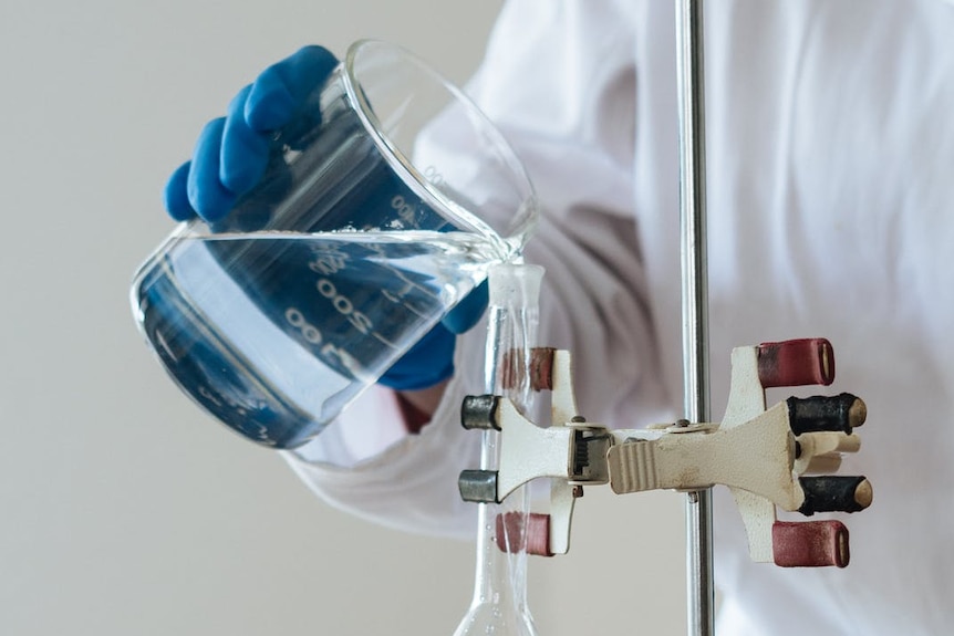 A person wearing blue gloves pours a clear liquid from a beaker into a test tube.