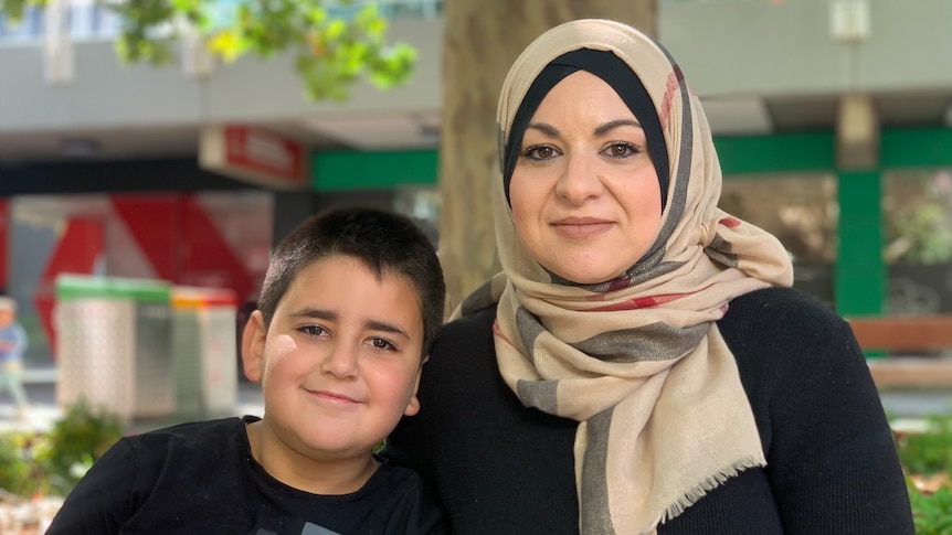 A young boy with a shaved head and black t-shirt leans into his mum, who wears a black top and cream headscarf.