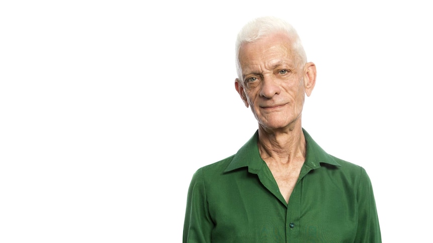 Robert Dessaix, a slim older white man with short white hair in a green shirt, stands looking at the camera.