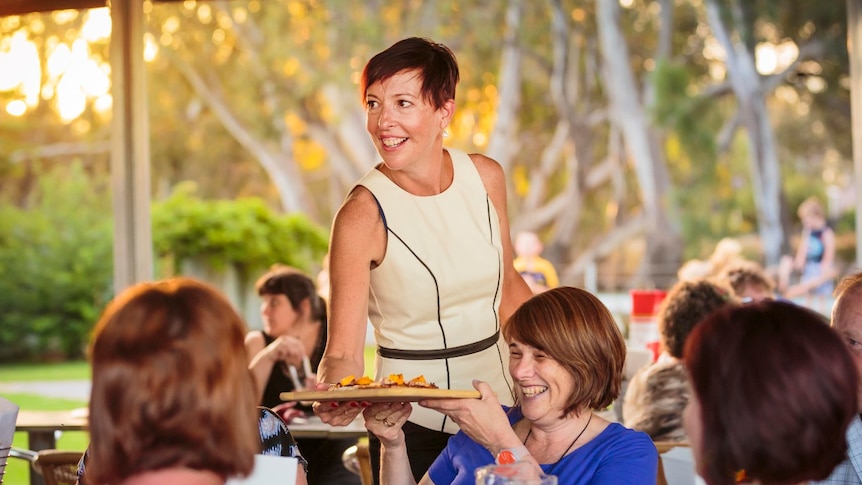 Lady delivers pizza to a table of diners in an outdoor dining area with trees and other diners in the background.