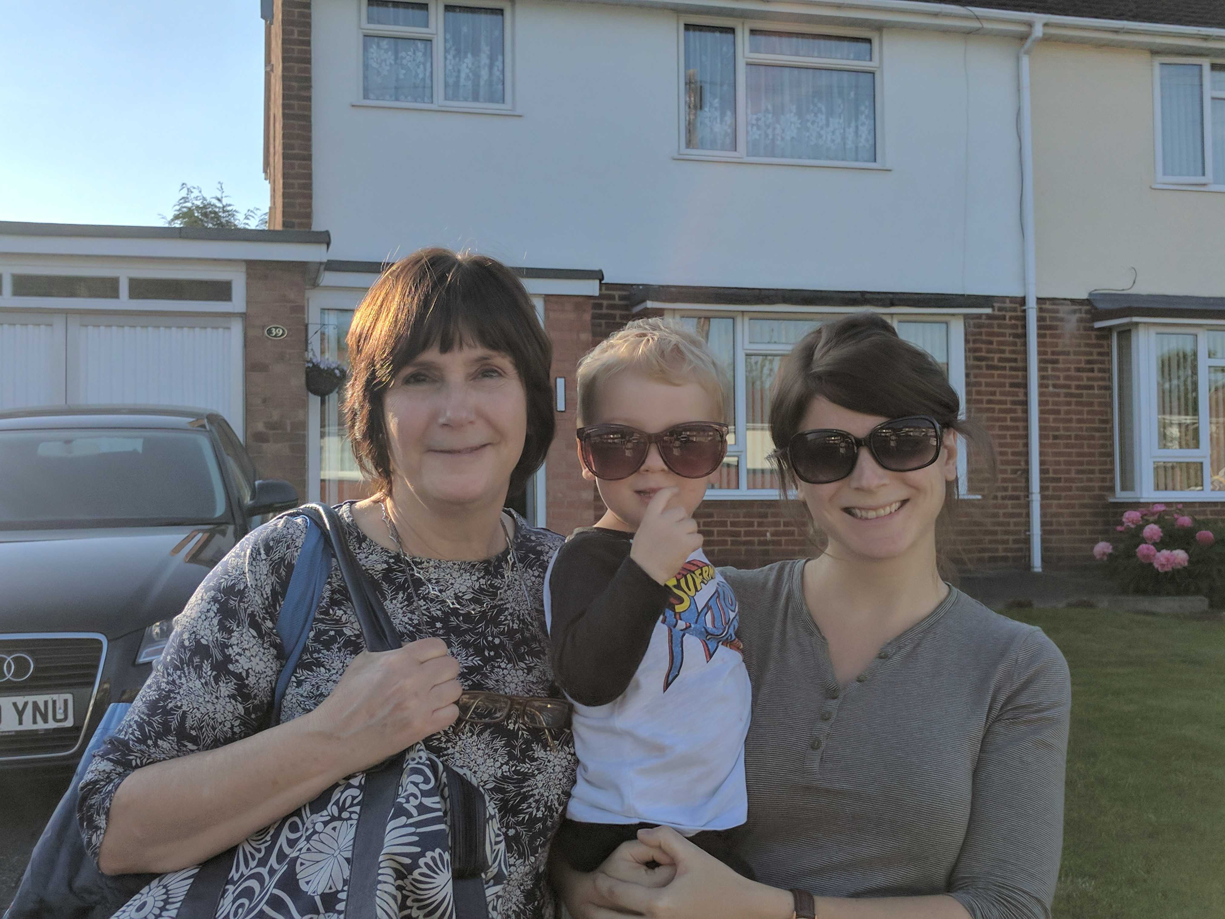 Woman holding her young son standing next to her mother outside a house.