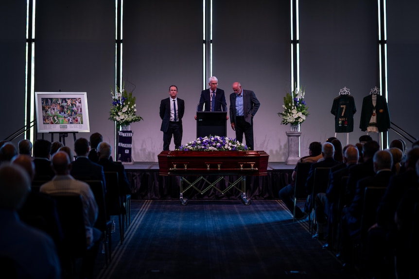 Three men stand on a stage behind a casket