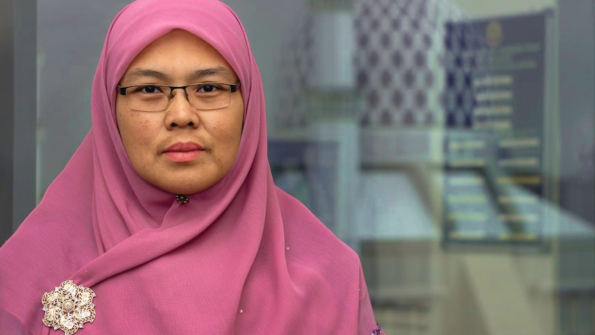 A woman wearing a pink hijab poses for a photo with a serious expression on her face.