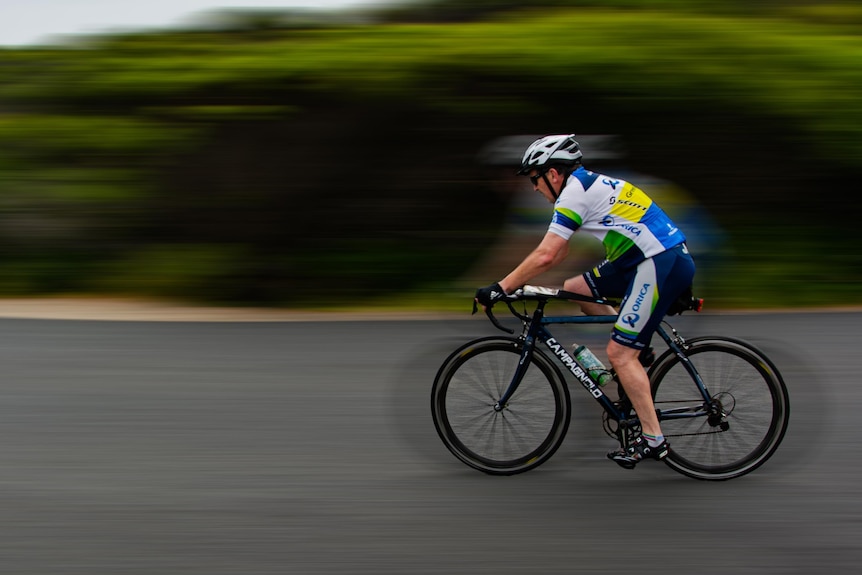 A man on a bike in lycra rides along a road flanked by green bushes.
