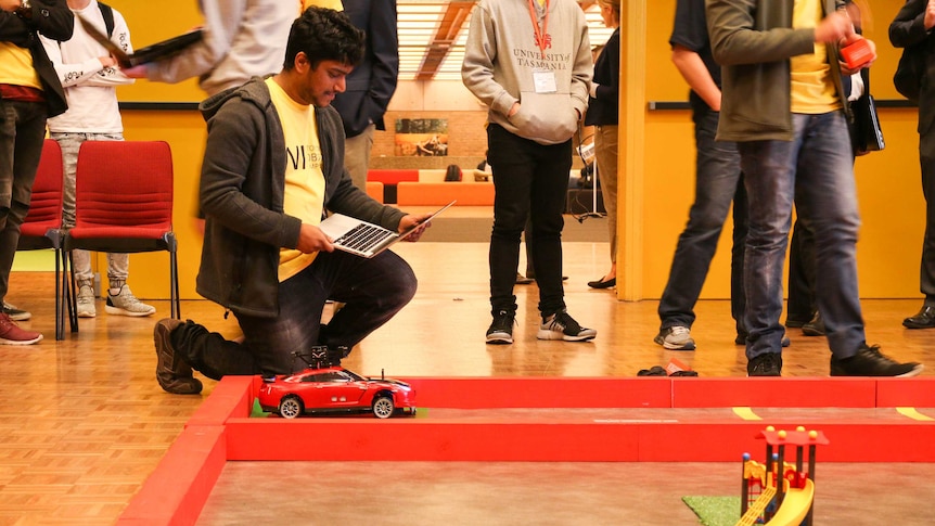 Uni student leans down beside a robot car holding a laptop