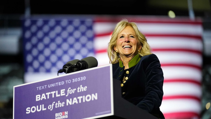 A woman with blond hair and wearing a long black coat stands in front of the US flag at a podium.