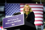 A woman with blond hair and wearing a long black coat stands in front of the US flag at a podium.