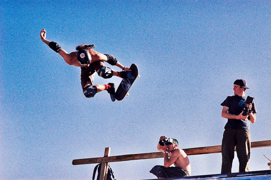 A photographer kneels to take a photo of a skateboarder doing a trick in the air.