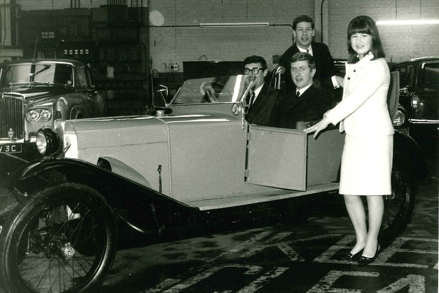 Three young men in a vintage car with a young woman opening the door