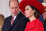 Prince William and Princess Catherine, wearing a suit and a red dress, stand waiting to welcome a dignitary