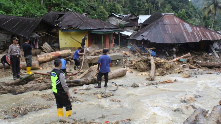 Rescue authorities stand surrounded by collapsed trees, damaged homes near jungle as water gushes on the ground.