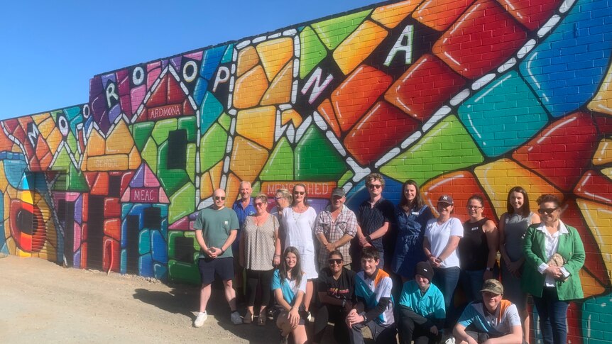 a group of people standing in front of an outdoor colourful mural on a brick wall in Mooroopna. 