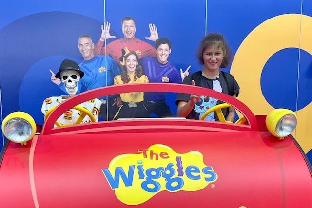 A skeleton wearing a black hat sits in a red Wiggles car beside a girl with brown hair.