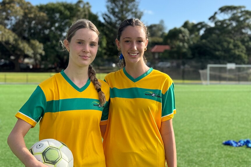 Two teenage girls wearing green and gold soccer jurnseys look at the camera, while one holds a soccer ball. 