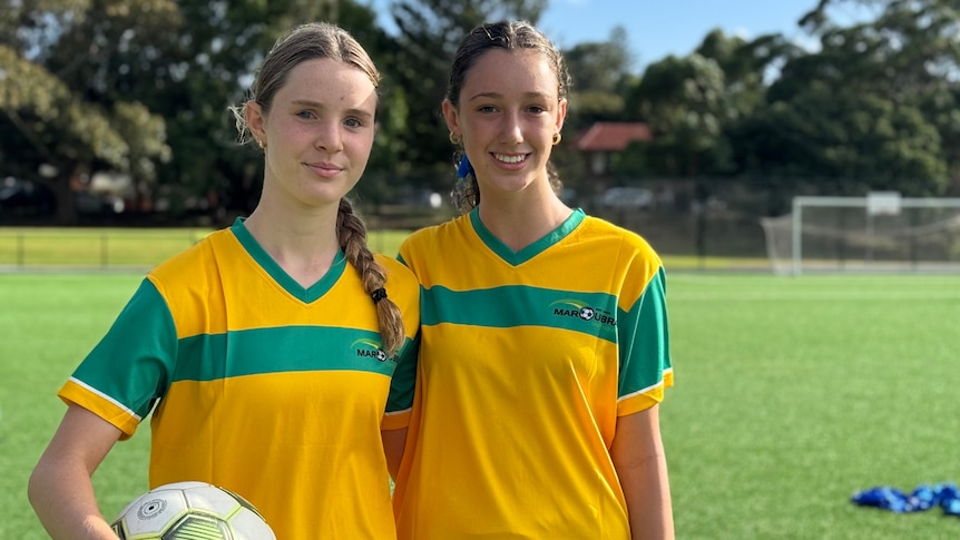 Two teenage girls wearing green and gold soccer jurnseys look at the camera, while one holds a soccer ball. 