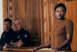 An African American woman stands in a courtroom dock, looking worried. Two white policeman glance in her direction.