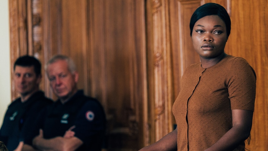 An African American woman stands in a courtroom dock, looking worried. Two white policeman glance in her direction.