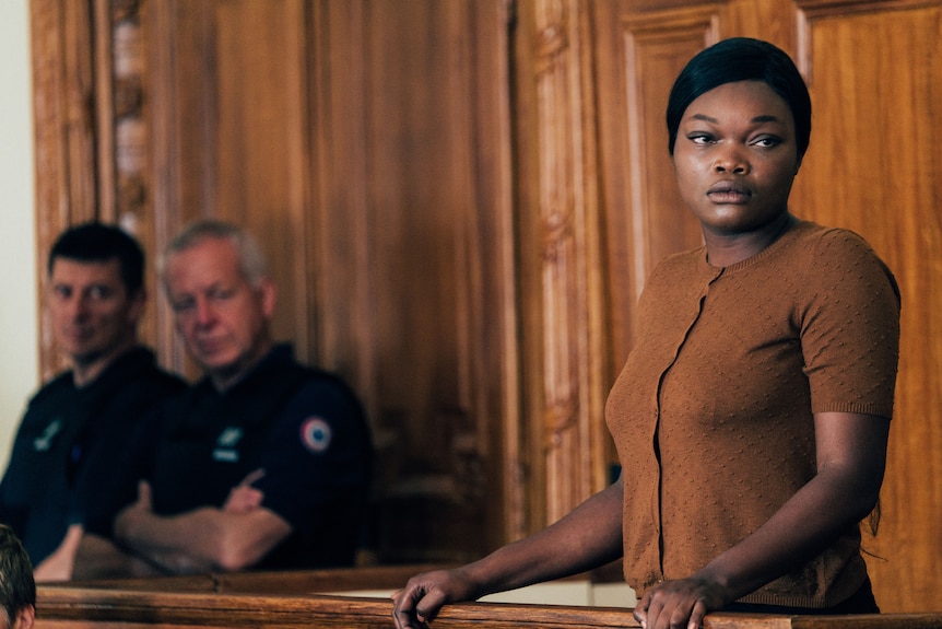 An African American woman stands in a courtroom dock, looking worried. Two white policeman glance in her direction.