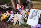 A framed photo of a young woman, bouquets of flowers and notes lie on the ground near logs.