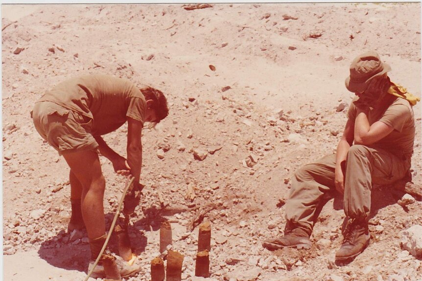 A soldiers sits on the ground watching his colleague pull some wire out of the ground.