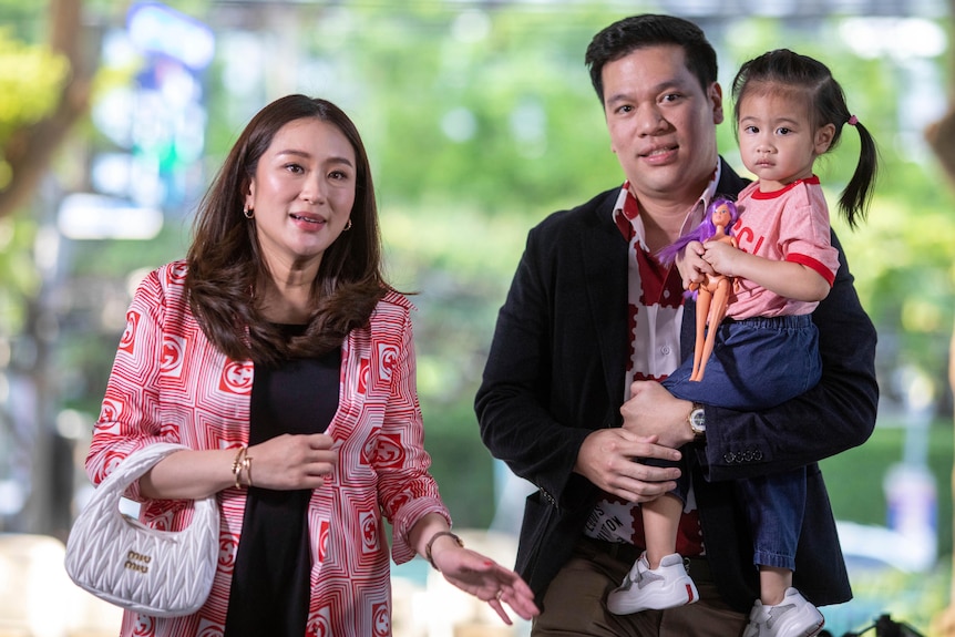 A Thai woman with long hair in red and white patterned blazer walks next to her husband and daughter.