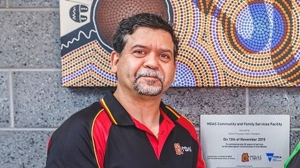 A man with a grey goatee wearing a red and black polo shirt in front of a plaque and Aboriginal artwork.