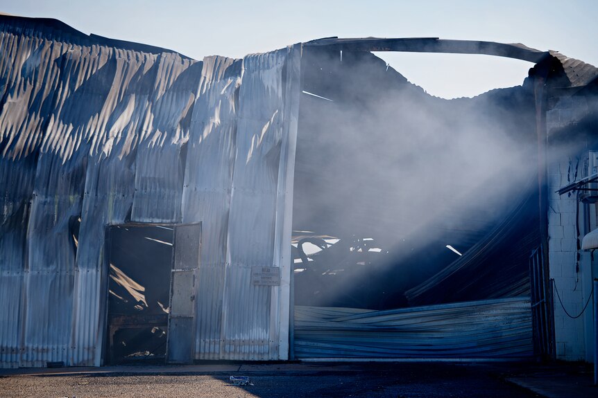 A large corrugated iron warehouse damaged by fire, with some sheets having fallen off and smoke lingering around it.