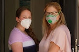 Robyn Cairnes and her daughter Ella stand arm in arm with face masks on in the doorway of a house