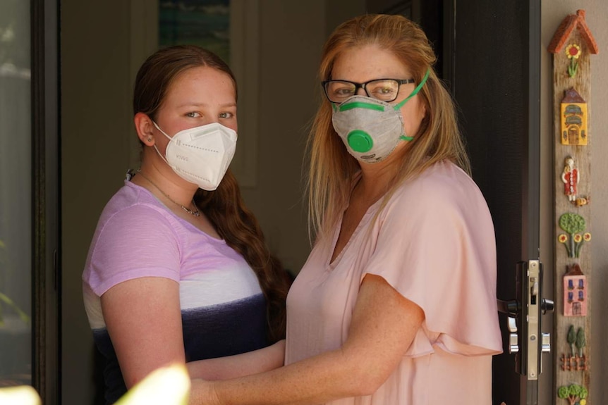 Robyn Cairnes and her daughter Ella stand arm in arm with face masks on in the doorway of a house