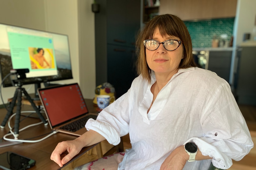 Dr Mitchell wears glasses and is dressed in a white top. She is sitting in front of a computer.