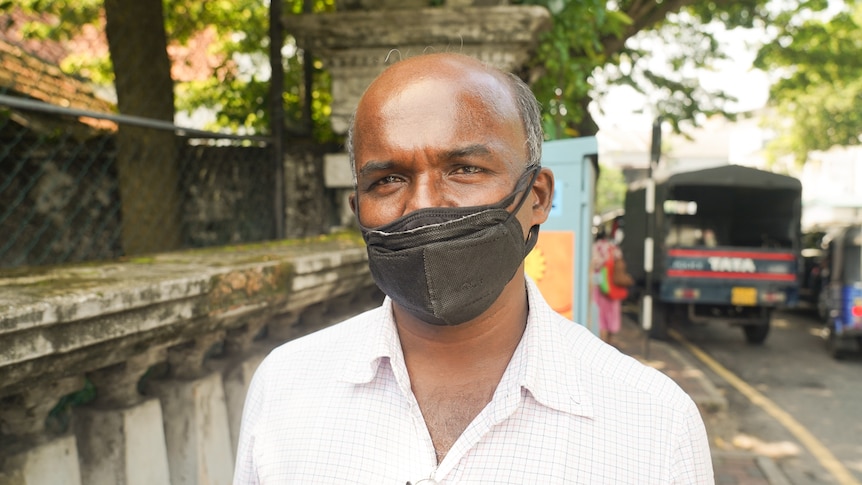 A black man wearing a face mask and white shirt stands near some trees.