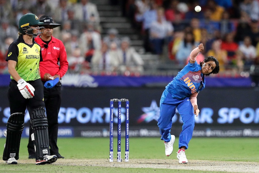 Poonam Yadav bowls a white cricket ball wearing blue cricket kit as an Australian batter stands next to her