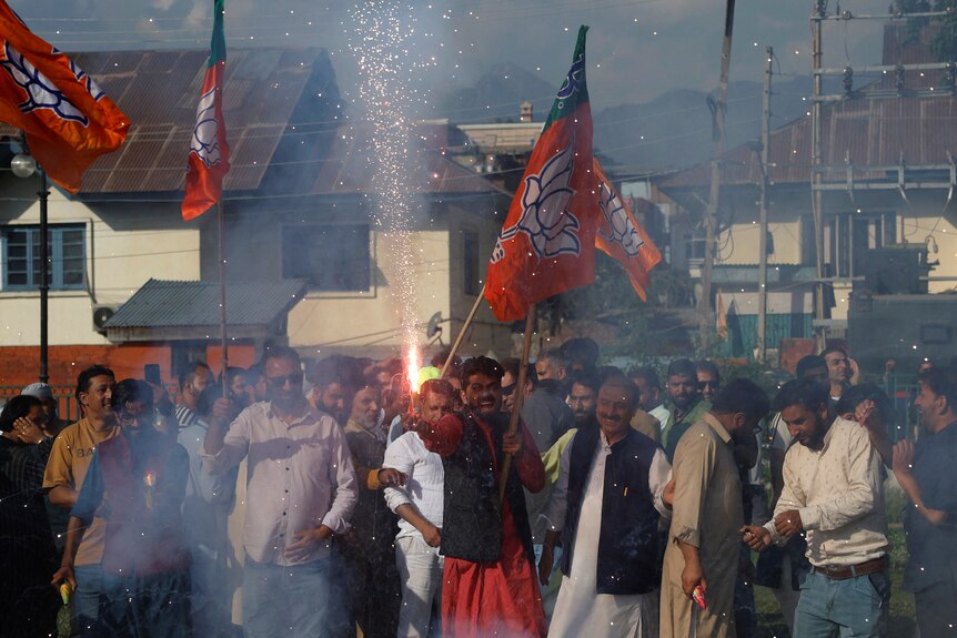 A man lets a firework off while holding a flag flying from a stick in the street surrounded by other people