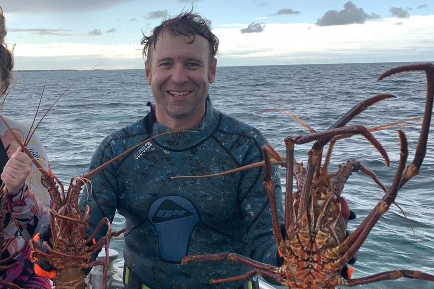 Smiling man on a boat wearing a wetsuit and holding two large rock lobsters.