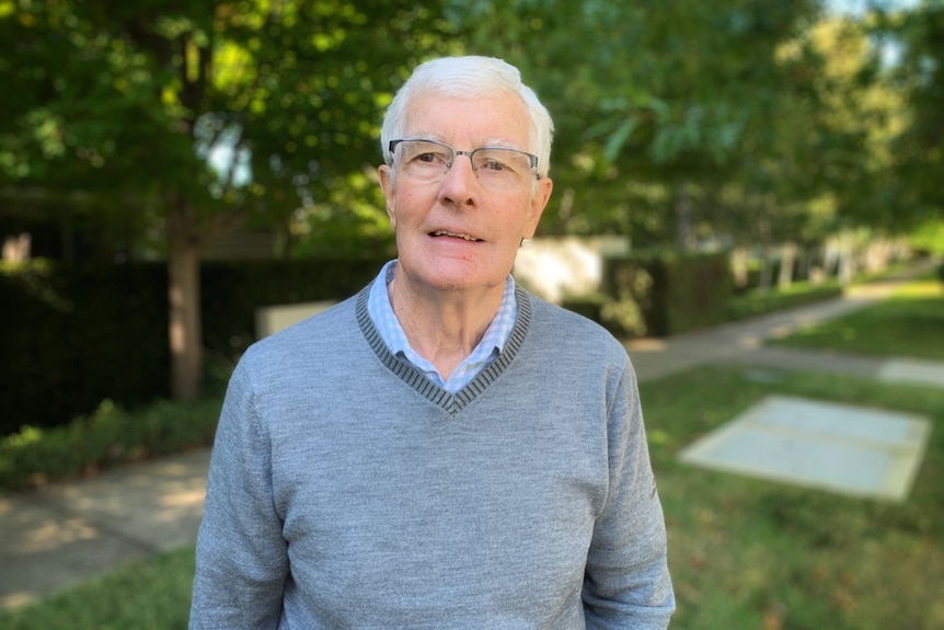 A man wearing a grey jumper with grey hair and glasses looks at the camera