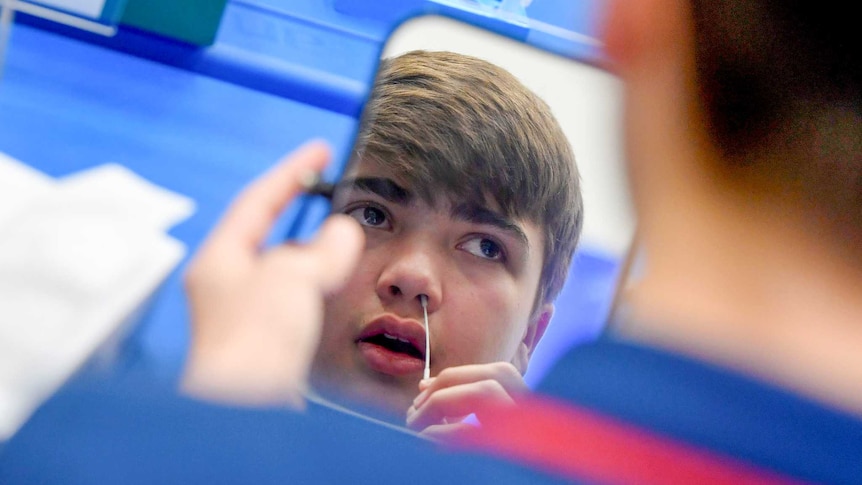 A teenaged boy holding a swab to his nose in front of a mirror