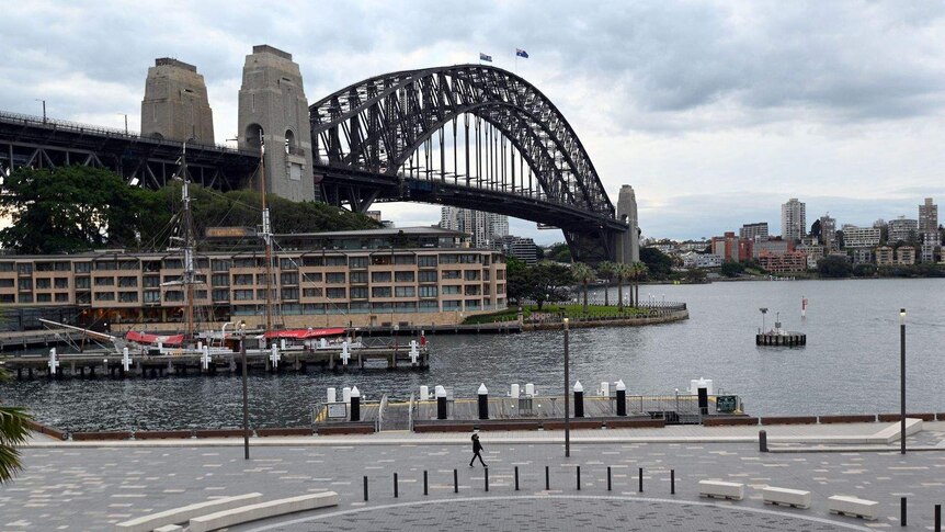A woman wearing a facemask strolls towards Sydney Opera House, the Harbour Bridge is in the background.