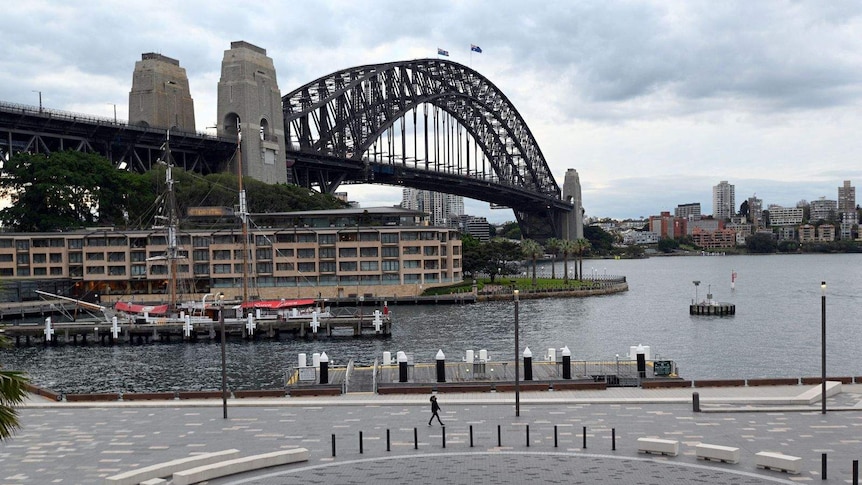 A woman wearing a facemask strolls towards Sydney Opera House, the Harbour Bridge is in the background.