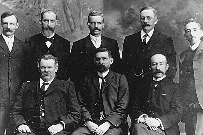 a black and white photo of nine men in early 20th century suits looking at the camera