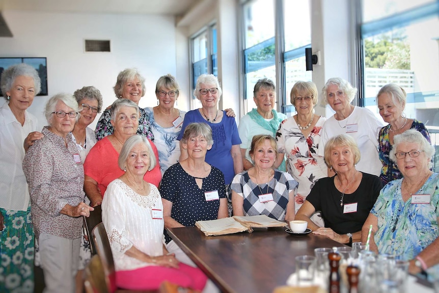 A group of older women stand posing for a photo indoors around an old book.