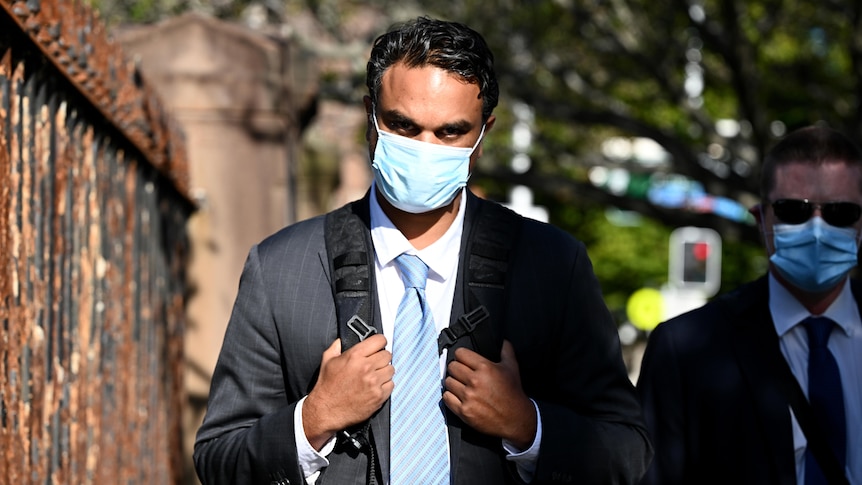 A male wearing a surgical mask walks past a metal fence holding the straps of his backpack